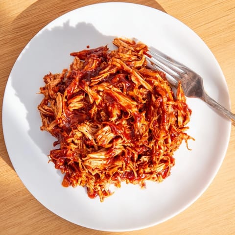 A close-up of saucy BBQ Pulled Chicken ready to be served alongside classic dill pickles.