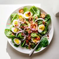 Spinach Salad with Bacon plated in a white bowl, featuring fresh spinach, crispy bacon crumbles, and sliced boiled eggs drizzled with tangy dressing.